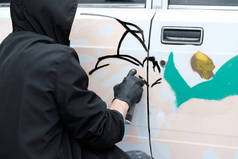 青年亚文化涂鸦。一个穿着黑色衣服和手套的家伙在一辆白色汽车上画了一个抽象的形象。破坏或艺术.