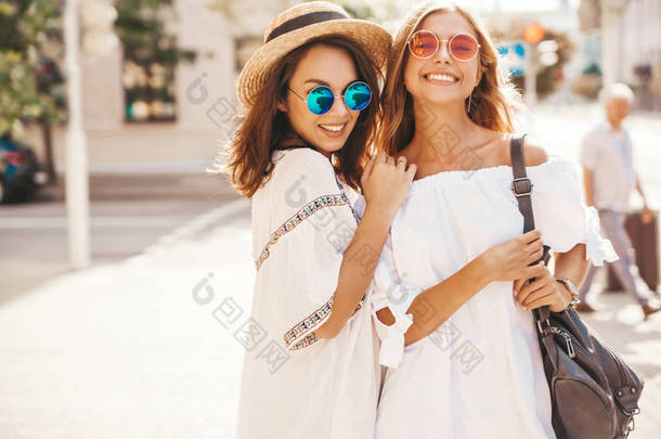 时尚肖像的两个年轻时尚的嬉皮黑发和金发女郎在夏季晴天穿着白色时髦的衣服摆在街头背景。疯了