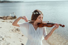 迷人的年轻女子优雅的礼服和花卉花圈在海边玩小提琴