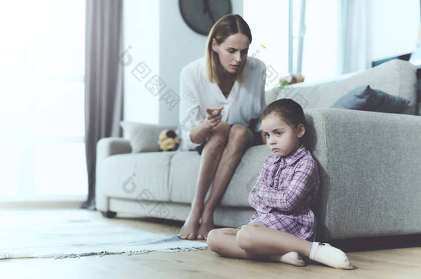 心烦意乱的女孩坐在地板上, 而母亲大喊大叫。小漂亮的孩子附近的光沙发采取不满, 当女人大喊大叫与分散的手。家庭关系概念