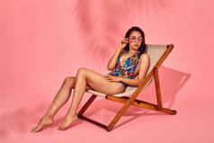 夏季生活方式的时尚肖像年轻惊艳的女人在躺椅上, 粉红色的背景, 工作室拍摄。穿着时尚的太阳镜, 比基尼泳装.