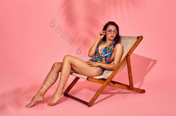 夏季生活方式的时尚肖像年轻惊艳的女人在躺椅上, 粉红色的背景, 工作室拍摄。穿着时尚的太阳镜, 比基尼泳装.