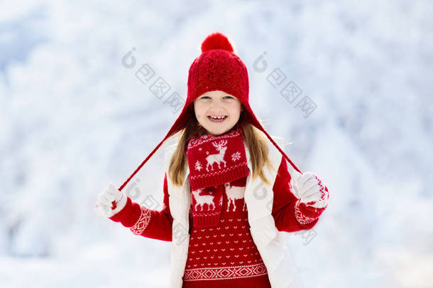 圣诞节假期,戴着红帽子的孩子在雪地里玩耍.冬天户外的乐趣。圣诞节前夕，孩子们在雪地公园玩耍。穿着针织毛衣、围巾和带圣诞装饰品的手套的小女孩. 