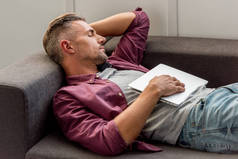 男子躺在沙发上与笔记本电脑和睡在家里的办公室
