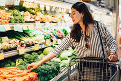 日本妇女在超市推购物车走在蔬菜和水果区。漂亮的亚洲女士拿起胡萝卜旁边花椰菜购买食品保健。妻子准备晚餐