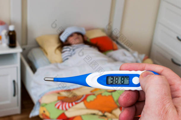 接近一个标有38摄氏度的数字温度计。小女孩躺在床上，额头上铺着湿布