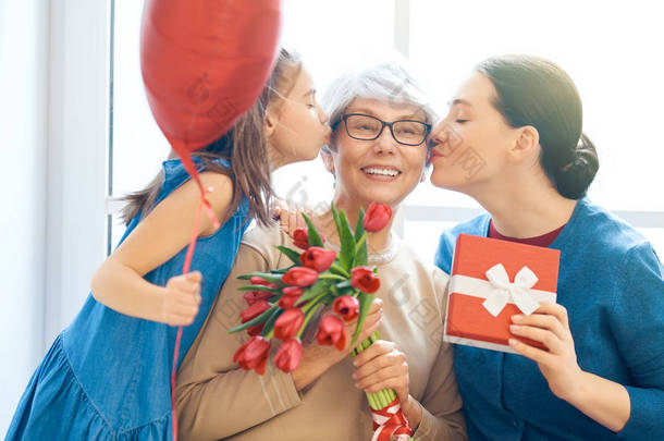 母亲节快乐！孩子的女儿在祝贺妈妈和奶奶送给他们的花郁金香。奶奶，妈妈和女孩笑着和拥抱。家庭假日和团聚.