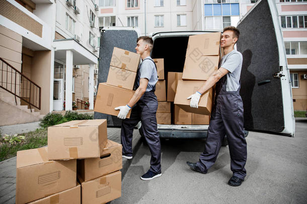 两个穿着制服的年轻英俊微笑的工人正在<strong>卸货</strong>, 面包车里<strong>装</strong>满了箱子。公寓楼在背景中。房子移动, 搬运工服务