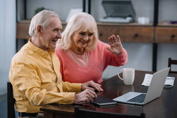 微笑的资深夫妇坐在桌子上, 并在家里的视频通话使用笔记本电脑