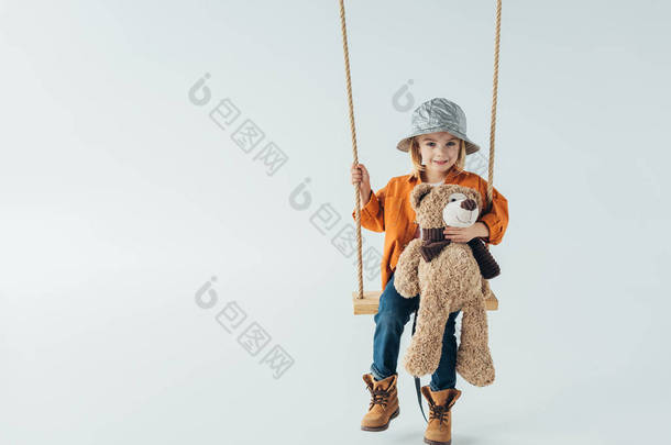 可爱的孩子在牛仔裤和橙色衬衫坐在秋千上, 抱着泰迪熊 