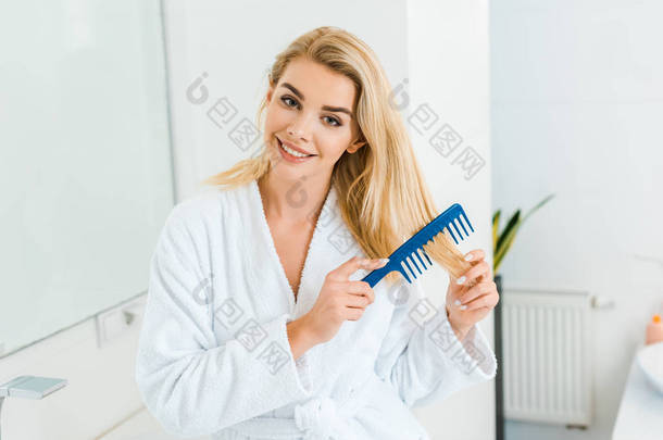 美丽和微笑的女人在白色浴衣看着相机, 并在浴室使用梳子 