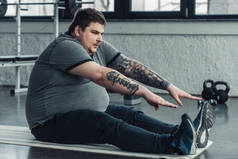 超重纹身男子坐在健身垫和伸展在健身房