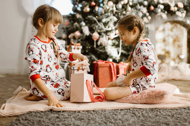 两个穿着睡衣的小姐妹坐在地毯上，在光线舒适的房间里打开<strong>新年礼</strong>物，还有美丽的<strong>新年</strong>树
