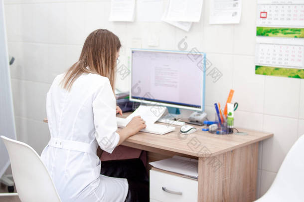 在诊所医院，白种金发女郎穿白色制服的女医生的背面视图在电脑上工作。背景柜和其他医疗设备。健康理念