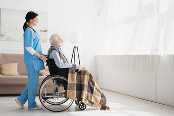 愉快的护士站在房间里与复制空间, 并携带约残疾的灰色头发男子在轮椅上的侧视图图片