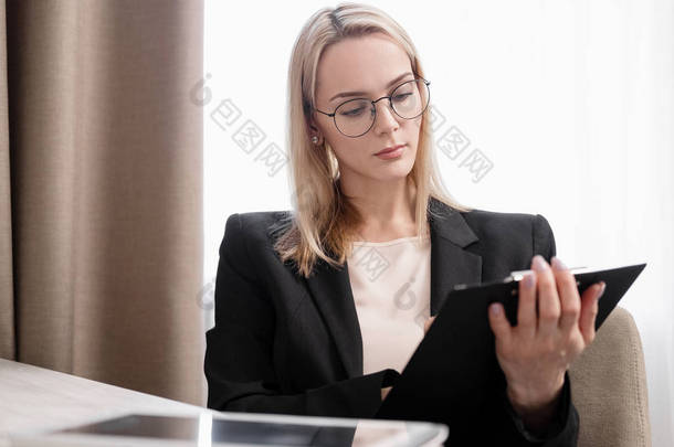 戴着眼镜、穿着西装的漂亮金发女郎在平板电脑上工作,在电话上交谈,在笔记本上做笔记。酒店房间,靠窗的桌子.