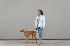 快乐的女孩和棕色的狗在灰色的墙壁背景。女人站在街上,看着远方,抱着一只宠物。复制空间