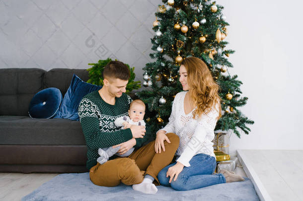 客厅里一个快乐的微笑和欢乐的家庭的肖像，装饰圣诞节。父亲抱着孩子，母亲看着他们，微笑着。家人坐在地板上。舒适的概念，庆祝在Chri新年