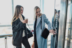 两个美丽的女孩，一个金发女郎和一个黑发女郎，穿着一件带窗户的玻璃电梯的外套。市中心的购物中心或办公室.