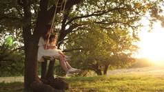 妈妈在阳光下摇摇着女儿在树下。母亲和婴儿骑在森林的橡树树枝上的绳索摆动。女孩笑了，高兴。家庭乐趣在公园，在自然。温暖的夏日.
