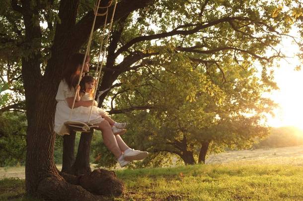 妈妈在阳光下摇摇着女儿在树下。母亲和婴儿骑在森林的橡树树枝上的绳索摆动。女孩笑了，高兴。家庭乐趣在公园，在自然。温暖的夏日.