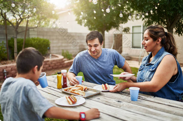 快乐的他的家人在后院的野餐桌上吃烤热狗