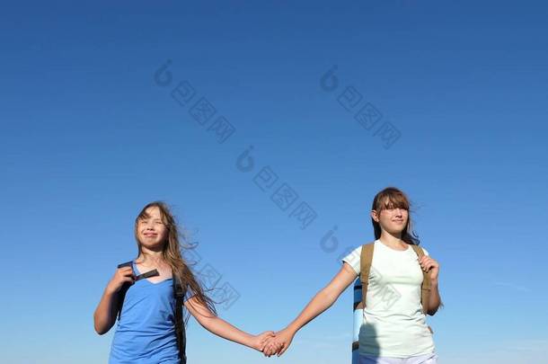 姑娘们，女旅行家手牵着手走在蓝天的背景上。 特写。 团队合作旅游青少年。 女旅行家走过田野。 夏天在草地上远足的姑娘们.