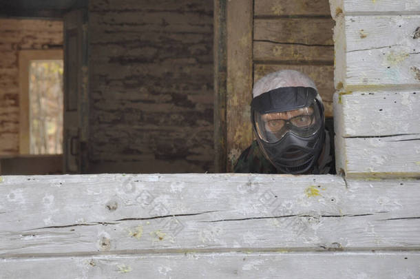 积极的彩弹游戏。 一位身穿防护服、戴着带有彩弹标志的面罩的彩弹手，正坐在一座老房子的埋伏中.