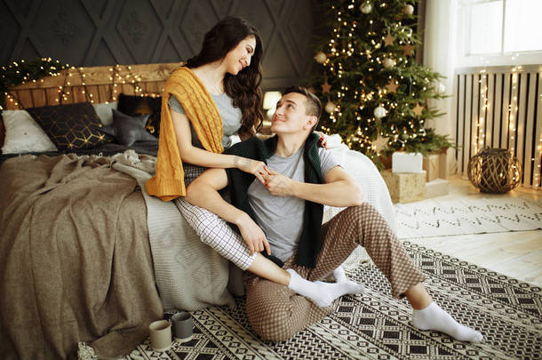 快乐的一对恋人在早晨的床上靠近圣诞树，拥抱着，圣诞节的早晨。 家居服和毛衣。 <strong>心灵</strong>的慰藉和温暖