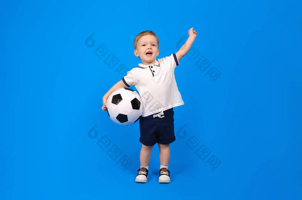 快乐的男孩微笑着站在足球旁边，为胜利而欢欣鼓舞，他的胳膊在蓝色的背景下高高地伸展着。胜利者和冠军的情感，放在文字上。赢得和实现目标的概念