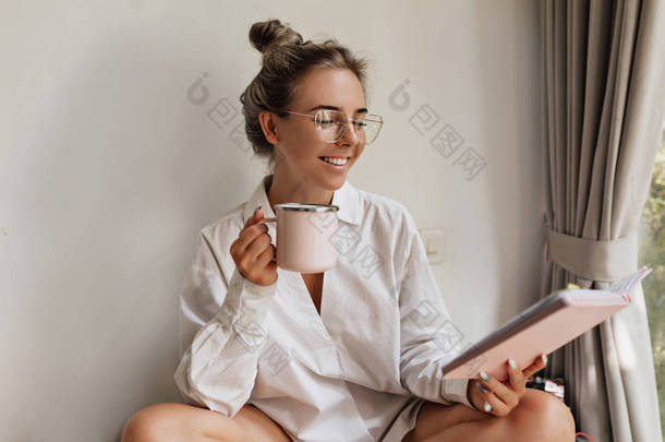 Happy smiling young woman with blond hair wearing spectaculars drinking coffee and reading notebook 