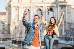 在城市的喷泉边，一个男人带着女朋友的笑容，拿着智能手机自拍