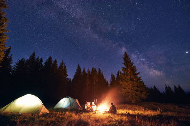 五人组的游客，男人和女人坐在灯火辉煌的帐篷边燃着篝火，享受着满天星斗的美丽夜空和灿烂的银河，温暖的夏夜。旅游业的概念