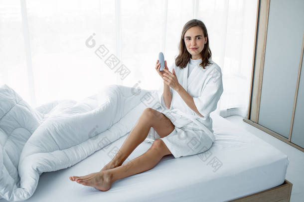 在家里的卧室里，一位漂亮的白人年轻女子在白色的床上用她的腿涂了些护肤霜。年轻女子在腿上涂抹奶油的抹布。美容美发的女人洗完澡也有护肤功能.