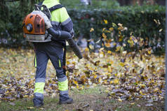 在城市公园操作重型吹叶机的工人。深蓝色和浅绿色的衣服。秋天摘下落叶。叶子在旋转.