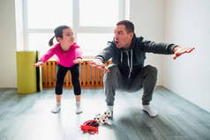 年轻的父亲和他可爱的小女儿正在家里做蹲姿练习。可爱的孩子和爸爸正在她房间窗户边的一个垫子上训练