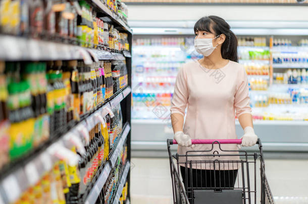 亚洲妇女，戴着卫生面具，戴着橡胶手套，购物车在杂货店里，正在寻找酱汁或调味品，准备在流感大流行期间进行检疫
