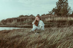 一位红润的年轻女子坐在湖边枯萎的草地上.一个红头发的女孩，穿着一件亚麻布的白色夹克和一条长裤，还有一件淡蓝色的衬衫。她正在享受一个好天气。复制空间.