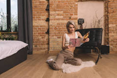 美丽的年轻女子坐在卧室里蓬松的地毯上，看杂志