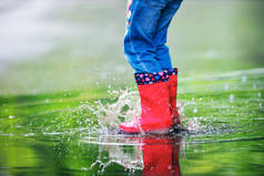 春天的一天，穿着红色橡胶靴的快乐小女孩在水坑里跳着走着.