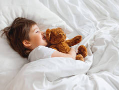 托德勒和可爱的泰迪熊躺在床上。在白色毛毯下的小男孩，毛绒玩具。刷子守卫着孩子们的睡眠.早上睡在舒适的家.