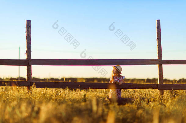 日落时分，戴帽子的女孩站在村子的篱笆旁边。走在农村.