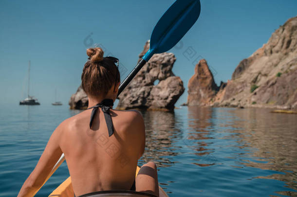一个穿着泳衣的女孩在一艘黄色的皮划艇上游泳，在海里划着蓝色的桨。在岩石和岩石海岸的背景下娱乐、<strong>旅游</strong>、<strong>旅游</strong>的概念。阳光灿烂的<strong>日</strong>子.