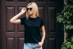 穿着黑色T恤，头戴眼镜，穿着时尚的金发女孩在街上摆姿势，穿着都市风格的服装。街头摄影