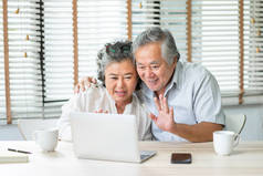 快乐的亚洲老夫老妻在家里用笔记本电脑与家人进行视频聊天和握手。微笑的老年男女喜欢在笔记本上使用互联网技术，喜欢成熟的生活方式