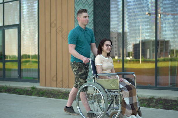 一个年轻的男人带着一个坐在轮椅上的年轻女人.一个助手载着一个<strong>残疾人</strong>坐在轮椅上沿街行走.<strong>残疾人</strong>特别交通.