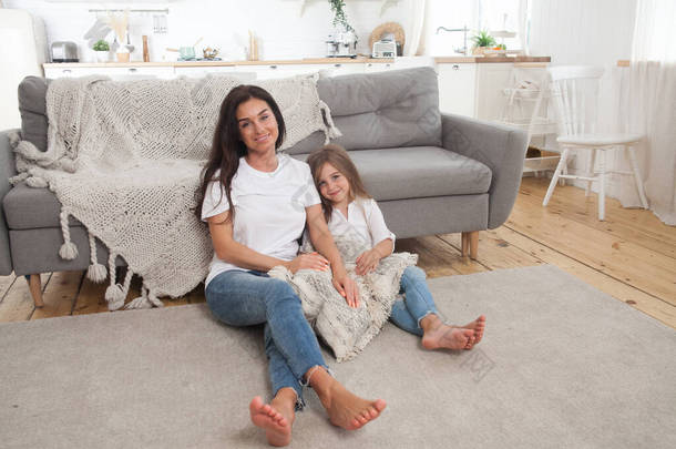 迷人的年轻妈妈和可爱的小女孩一起呆在家里，坐在沙发旁边，铺着灰色舒适的毯子。快乐的家庭斯堪的纳维亚<strong>风格客厅</strong>内部.
