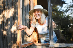 快乐的微笑着的老妇人，穿着草帽，站在开着的窗前，摆出一副很开心的样子。退休的老年人的概念。乡间别墅的检疫
