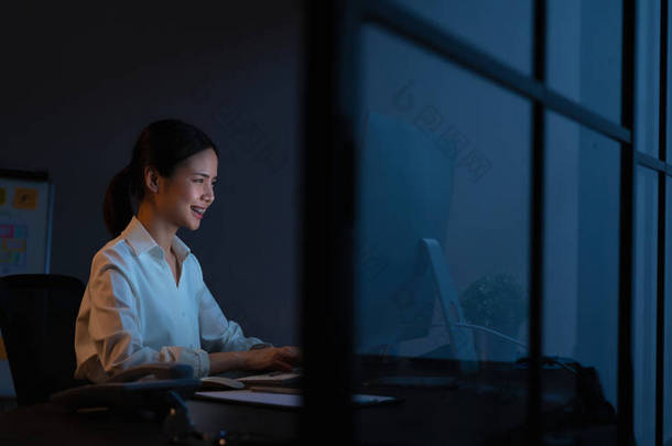 微笑着坐在桌旁，晚上在办公室的电脑上工作的亚洲女商人.