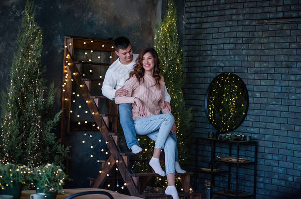 恋爱中的幸福夫妻坐在一个装饰着彩灯和树木装饰的手工制作的木制楼梯上。一个年轻人从背后拥抱着一个漂亮的卷曲女孩，他们笑着，亲吻着期待着圣诞节的到来